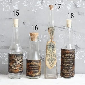 Puede incluir: Cuatro botellas de vidrio transparente con tapones de corcho, numeradas 15, 16, 17 y 18. Las botellas 15, 16 y 18 tienen etiquetas de papel rectangulares oscuras de estilo antiguo con texto. La 17 tiene un cuello largo y fino y una etiqueta floral vertical.