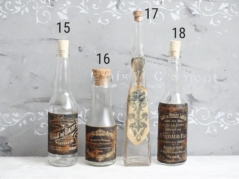 Puede incluir: Cuatro botellas de vidrio transparente con tapones de corcho, numeradas 15, 16, 17 y 18. Las botellas 15, 16 y 18 tienen etiquetas de papel rectangulares oscuras de estilo antiguo con texto. La 17 tiene un cuello largo y fino y una etiqueta floral vertical.