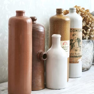 Könnte beinhalten: Eine Sammlung antiker Keramikflaschen in verschiedenen Braun- und Weißtönen. Die Flaschen haben unterschiedliche Formen und Größen, einige mit Etiketten. Die Anordnung umfasst eine hohe, zylindrische Flasche und eine weiße, abgerundete Flasche.