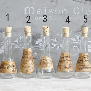 Puede incluir: Cinco botellas de vidrio transparente con cuellos estrechos y tapones de corcho, cada una con una etiqueta de papel beige con texto negro. Las botellas est&aacute;n numeradas del uno al cinco en negro sobre cada art&iacute;culo en una fila.