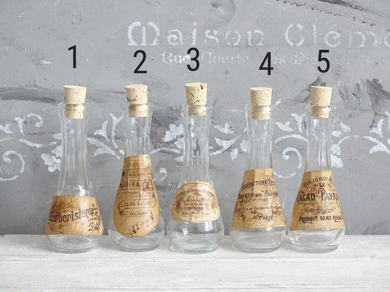 Puede incluir: Cinco botellas de vidrio transparente con cuellos estrechos y tapones de corcho, cada una con una etiqueta de papel beige con texto negro. Las botellas est&aacute;n numeradas del uno al cinco en negro sobre cada art&iacute;culo en una fila.