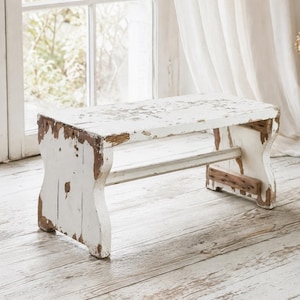 Puede incluir: Un banco rectangular de madera con un acabado de pintura blanca desgastada que revela la madera marrón natural debajo. El banco tiene dos soportes laterales sólidos conectados por una barra transversal, mostrando una textura envejecida y gastada.