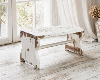 Peut inclure: Un banc rectangulaire en bois avec une finition peinte en blanc vieilli qui révèle le bois brun naturel en dessous. Le banc possède deux supports latéraux solides reliés par une barre transversale, présentant une texture usée et patinée.
