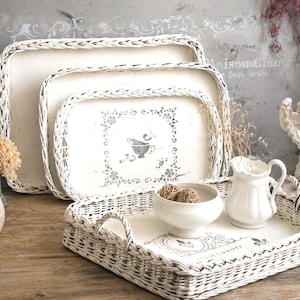 Op de afbeelding: Een set van drie rechthoekige witte rieten dienbladen met gevlochten randen en platte bodems. De dienbladen hebben een verweerde witte afwerking met grijze sjabloonmotieven van bloemen en kopjes op de binnenzijde en twee geïntegreerde handvatten.