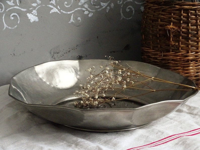 May include: A silver serving bowl with a scalloped edge, sitting on a white linen tablecloth with a red stripe. A sprig of dried white flowers is resting in the bowl.