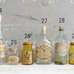 Puede incluir: Una colecci&oacute;n de seis botellas y frascos de vidrio con tapones de corcho, numerados del 24 al 29. Los recipientes var&iacute;an en forma, tama&ntilde;o y color, incluyendo vidrio transparente, &aacute;mbar y verde claro, con etiquetas de papel vintage.
