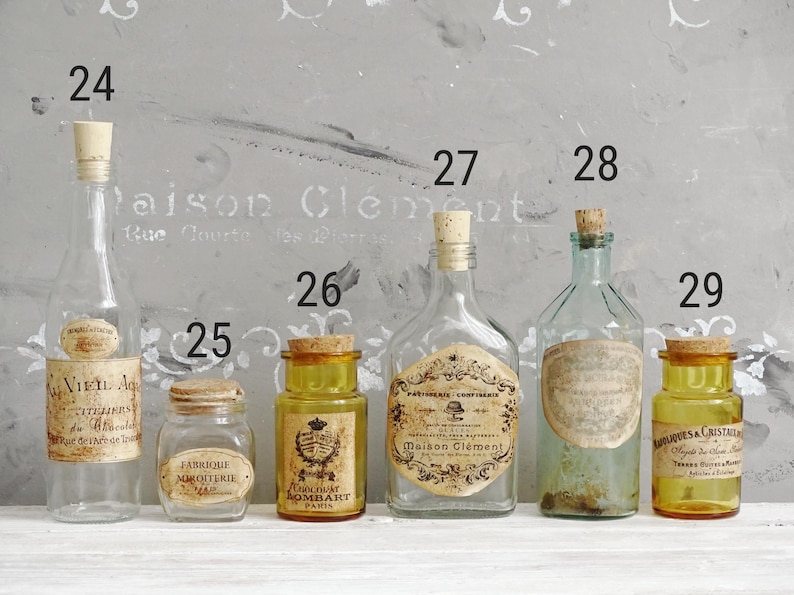 Puede incluir: Una colecci&oacute;n de seis botellas y frascos de vidrio con tapones de corcho, numerados del 24 al 29. Los recipientes var&iacute;an en forma, tama&ntilde;o y color, incluyendo vidrio transparente, &aacute;mbar y verde claro, con etiquetas de papel vintage.