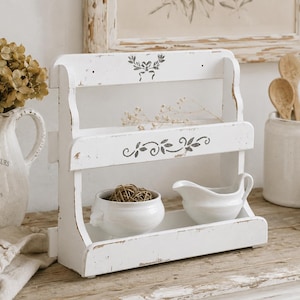 May include: A white wooden two-tier shelf with a distressed finish and gray stenciled floral motifs. The shelf features an open design with two horizontal surfaces, currently holding a small white ceramic bowl and a white ceramic gravy boat.