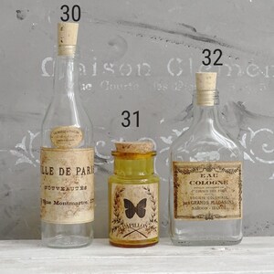 Puede incluir: Tres botellas de vidrio con tapones de corcho numeradas 30, 31 y 32. La botella 30 es alta y transparente con una etiqueta Ville de Paris. El frasco 31 es amarillo con un gr&aacute;fico de mariposa y la palabra Papillon. La botella 32 tiene una etiqueta de Eau de Cologne.
