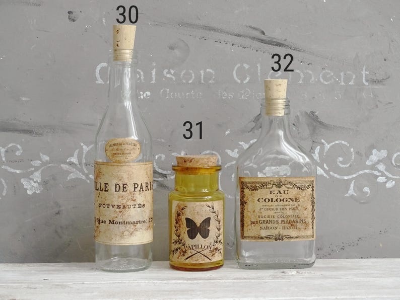 Puede incluir: Tres botellas de vidrio con tapones de corcho numeradas 30, 31 y 32. La botella 30 es alta y transparente con una etiqueta Ville de Paris. El frasco 31 es amarillo con un gr&aacute;fico de mariposa y la palabra Papillon. La botella 32 tiene una etiqueta de Eau de Cologne.