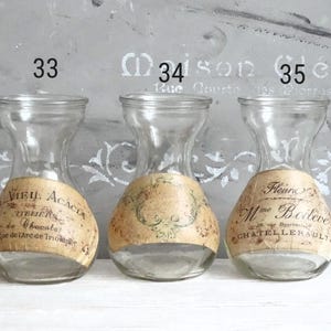 Puede incluir: Tres jarrones de vidrio transparente con cuello estrecho y base ancha, cada uno con una etiqueta de papel beige con tipograf&iacute;a negra de estilo antiguo y adornos decorativos. Los jarrones est&aacute;n numerados 33, 34 y 35 arriba.