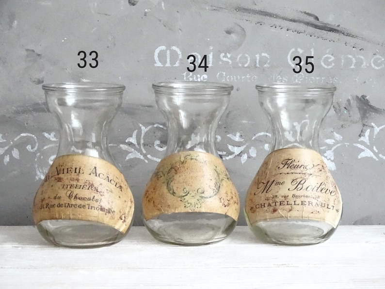 Puede incluir: Tres jarrones de vidrio transparente con cuello estrecho y base ancha, cada uno con una etiqueta de papel beige con tipograf&iacute;a negra de estilo antiguo y adornos decorativos. Los jarrones est&aacute;n numerados 33, 34 y 35 arriba.