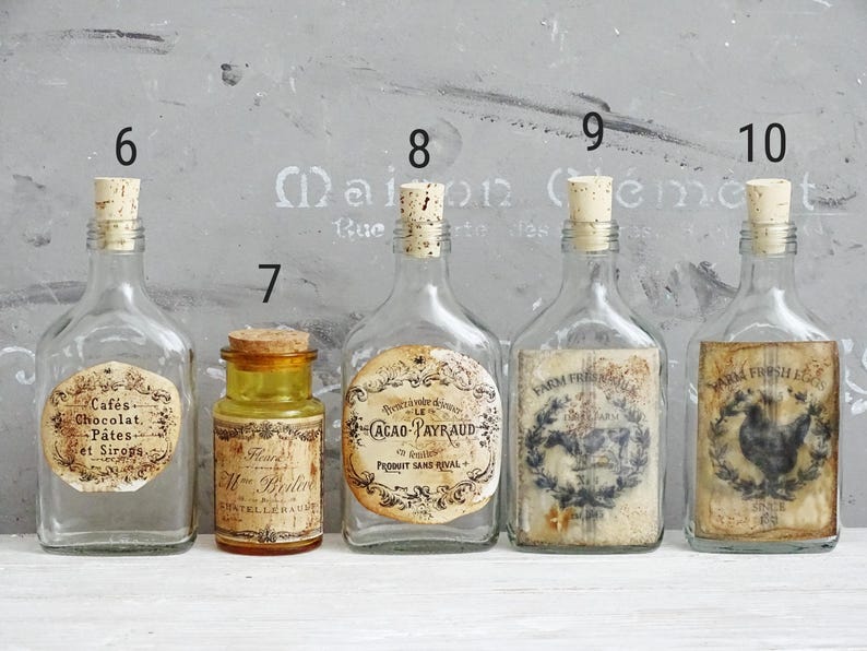 Puede incluir: Cinco botellas de vidrio numeradas 6, 7, 8, 9 y 10. Cuatro son de vidrio transparente con tapones de corcho y etiquetas de papel estilo vintage. Una es un frasco de vidrio &aacute;mbar m&aacute;s peque&ntilde;o con tap&oacute;n de corcho y etiqueta con texto.