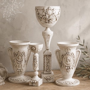 Puede incluir: Un conjunto de cuatro recipientes y pedestales decorativos de color blanco roto con intrincados relieves de escudos, coronas de laurel y motivos clásicos. El set incluye dos jarrones en forma de urna, un cáliz alto y un portavelas esbelto.