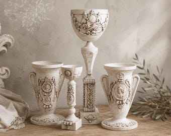 Può includere: Un set di quattro vasi e piedistalli decorativi color bianco sporco con intricati rilievi raffiguranti stemmi, ghirlande di alloro e motivi classici. Il set comprende due vasi a forma di urna, un calice alto e un portacandele slanciato.