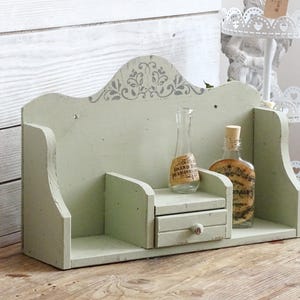 May include: A distressed, light green wooden shelf with a decorative top and a small drawer. Two glass bottles with cork stoppers and labels are displayed on the shelf. The shelf is on a wooden surface.