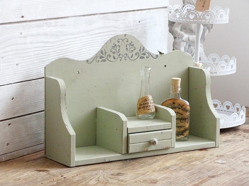 May include: A distressed, light green wooden shelf with a decorative top and a small drawer. Two glass bottles with cork stoppers and labels are displayed on the shelf. The shelf is on a wooden surface.