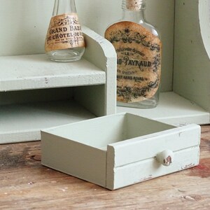 May include: A distressed, light green wooden shelf unit with a small drawer and two glass bottles with vintage labels. The drawer has a small, round knob. The unit is on a wooden surface.
