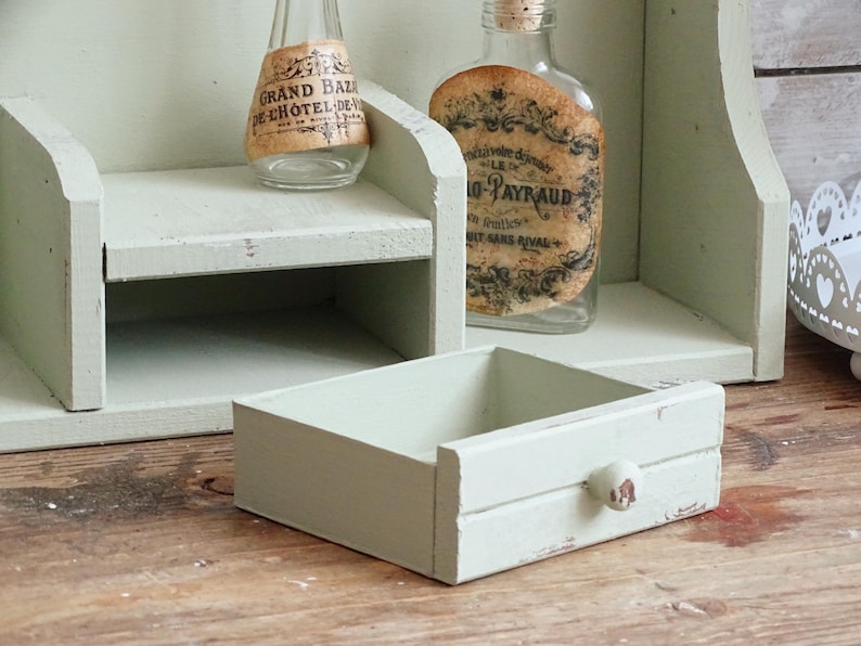 May include: A distressed, light green wooden shelf unit with a small drawer and two glass bottles with vintage labels. The drawer has a small, round knob. The unit is on a wooden surface.