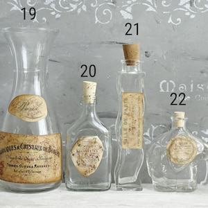 Puede incluir: Cuatro botellas de vidrio transparente numeradas 19, 20, 21 y 22. La 19 es una jarra ancha, la 20 es cuadrada, la 21 es alta y ondulada, y la 22 es redonda con asa. Cada una tiene una etiqueta de papel de estilo antiguo con texto.