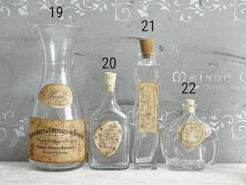 Puede incluir: Cuatro botellas de vidrio transparente numeradas 19, 20, 21 y 22. La 19 es una jarra ancha, la 20 es cuadrada, la 21 es alta y ondulada, y la 22 es redonda con asa. Cada una tiene una etiqueta de papel de estilo antiguo con texto.