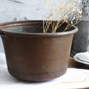 May include: A large, copper pot with a handle. The pot is old and has a patina of age. It is sitting on a white surface.