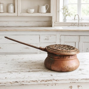 Puede incluir: Una olla redonda de cobre con un mango de metal largo y delgado y una tapa circular perforada extra&iacute;ble. La superficie de cobre muestra signos de oxidaci&oacute;n y desgaste, con un patr&oacute;n decorativo texturizado alrededor del borde de la tapa.