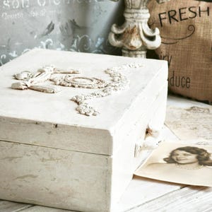 May include: A white, distressed wooden box with ornate, raised relief detailing on the lid. The box has a clasp and a small handle. A vintage photograph and a burlap pillow with the word "FRESH" are in the background.