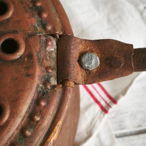Puede incluir: Un objeto de cobre desgastado con una parte superior circular que presenta remaches elevados y m&uacute;ltiples aberturas redondas. Un mango de metal plano y oxidado est&aacute; unido al lateral con un remache texturizado, mostrando signos de antig&uuml;edad.