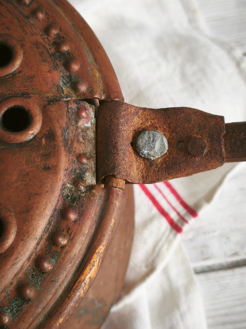Puede incluir: Un objeto de cobre desgastado con una parte superior circular que presenta remaches elevados y m&uacute;ltiples aberturas redondas. Un mango de metal plano y oxidado est&aacute; unido al lateral con un remache texturizado, mostrando signos de antig&uuml;edad.