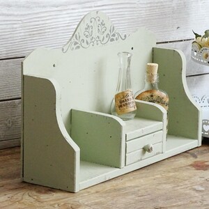 May include: A distressed, light green wooden shelf with a small drawer and decorative top. Two glass bottles with cork stoppers sit on the shelf. One bottle has a label that reads "GRAND DE CHOIX".