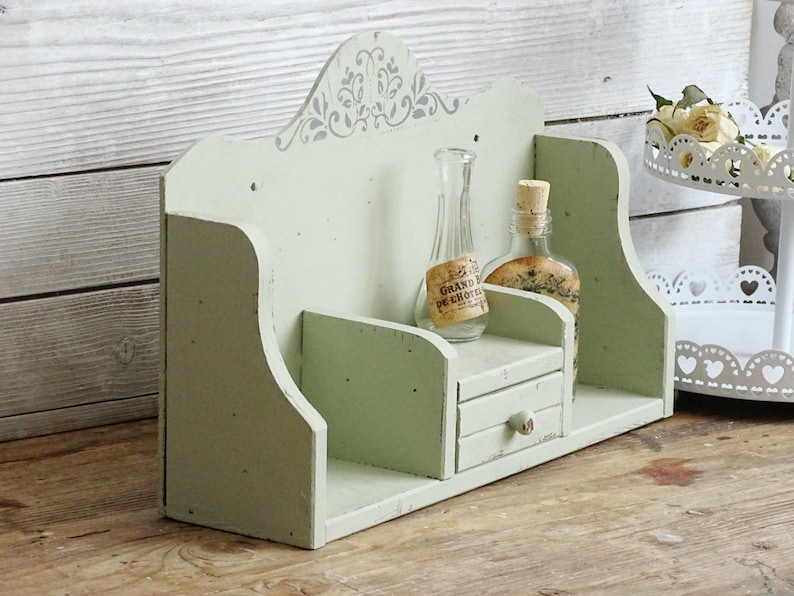 May include: A distressed, light green wooden shelf with a small drawer and decorative top. Two glass bottles with cork stoppers sit on the shelf. One bottle has a label that reads "GRAND DE CHOIX".