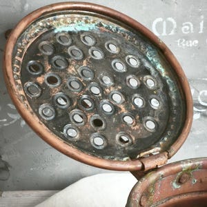 Puede incluir: Una tapa redonda de cobre con dise&ntilde;o de bisagra que presenta una superficie perforada con numerosos orificios circulares. El metal muestra signos de oxidaci&oacute;n con p&aacute;tina verde visible y &aacute;reas oscuras y desgastadas en la superficie.