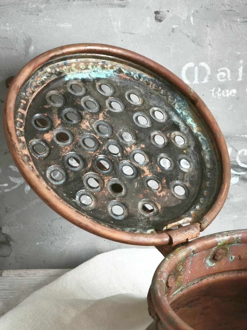 Puede incluir: Una tapa redonda de cobre con dise&ntilde;o de bisagra que presenta una superficie perforada con numerosos orificios circulares. El metal muestra signos de oxidaci&oacute;n con p&aacute;tina verde visible y &aacute;reas oscuras y desgastadas en la superficie.