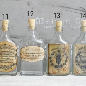 Puede incluir: Cuatro botellas de vidrio transparente con tapones de corcho, numeradas del 11 al 14. La botella 11 tiene una etiqueta redonda con el texto Senez-Sturbelle. La 12 tiene una etiqueta que dice Visiter Ville de Paris. La 13 muestra una abeja y Fresh Honey. La 14 muestra una oveja y Sheep Wool Farm.