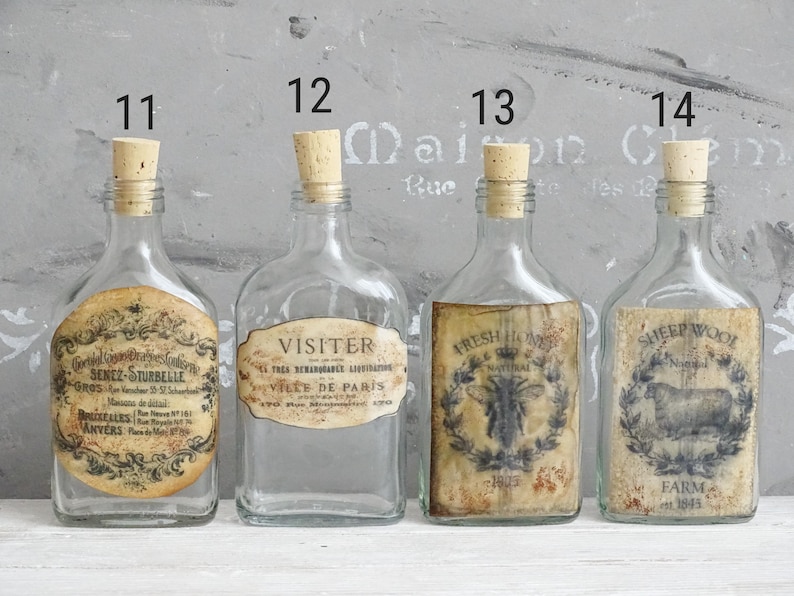 Puede incluir: Cuatro botellas de vidrio transparente con tapones de corcho, numeradas del 11 al 14. La botella 11 tiene una etiqueta redonda con el texto Senez-Sturbelle. La 12 tiene una etiqueta que dice Visiter Ville de Paris. La 13 muestra una abeja y Fresh Honey. La 14 muestra una oveja y Sheep Wool Farm.