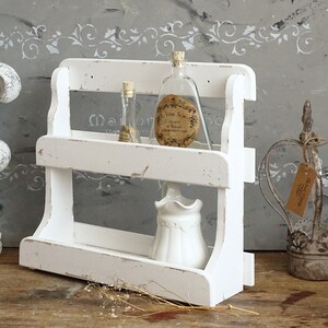 May include: A two-tiered white wooden shelf with a distressed finish. The top shelf holds two glass bottles with cork stoppers and labels, while the bottom shelf contains a small white ceramic pitcher. The shelf has curved side supports.