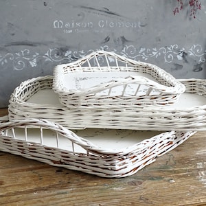 Op de afbeelding: Drie witte rieten dienbladen met een decoratief ontwerp. De dienbladen zijn op elkaar gestapeld. Het bovenste dienblad heeft een verhoogde rand en een decoratief ontwerp op de bodem.