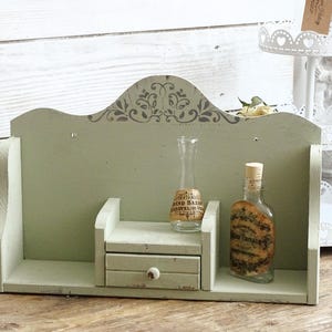 May include: A distressed, sage green wooden shelf with a small drawer and two glass bottles. The shelf has a decorative floral design at the top. One bottle has a cork stopper and a label. The other bottle has a cork stopper and a label.