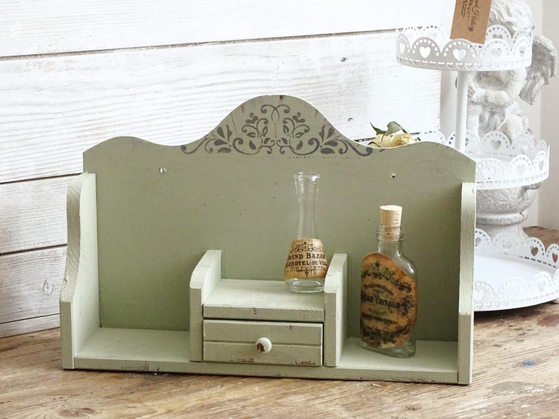 May include: A distressed, sage green wooden shelf with a small drawer and two glass bottles. The shelf has a decorative floral design at the top. One bottle has a cork stopper and a label. The other bottle has a cork stopper and a label.