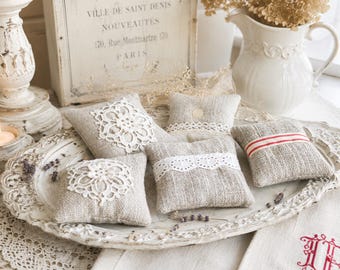 May include: A collection of four small, square, beige linen sachets arranged on an ornate, oval, white decorative tray. Two sachets feature white crocheted lace doilies, one has a white lace trim, and one displays a single horizontal red stripe.