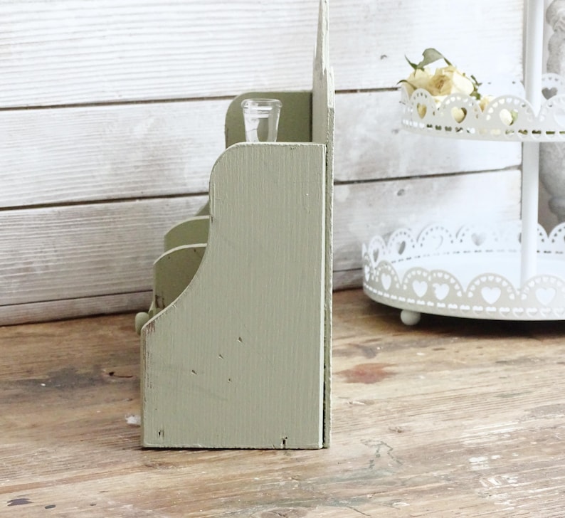 May include: A distressed, sage green wooden organizer with multiple compartments. A small, clear glass container sits in the top section. The organizer has a rustic, vintage aesthetic, perfect for home decor or storage.