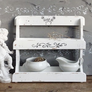 May include: A two-tiered white wooden shelf with a distressed finish and gray stenciled floral motifs. The shelf holds a small white ceramic bowl containing a woven sphere and a white ceramic gravy boat with a handle on the right side.
