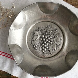 May include: A silver metal serving dish with an embossed design of grapes and leaves. The dish has a scalloped edge and a hammered finish.