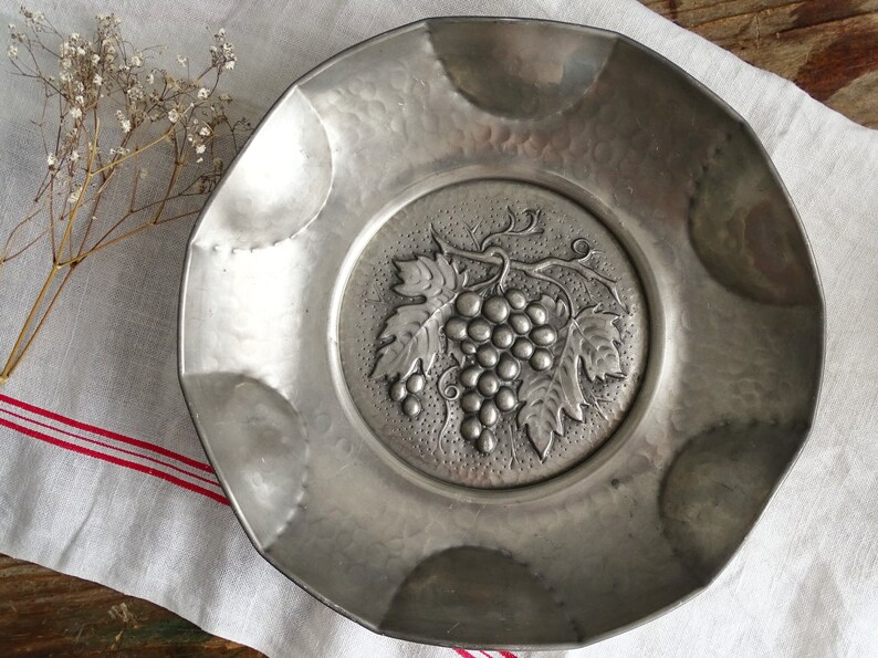 May include: A silver metal serving dish with an embossed design of grapes and leaves. The dish has a scalloped edge and a hammered finish.