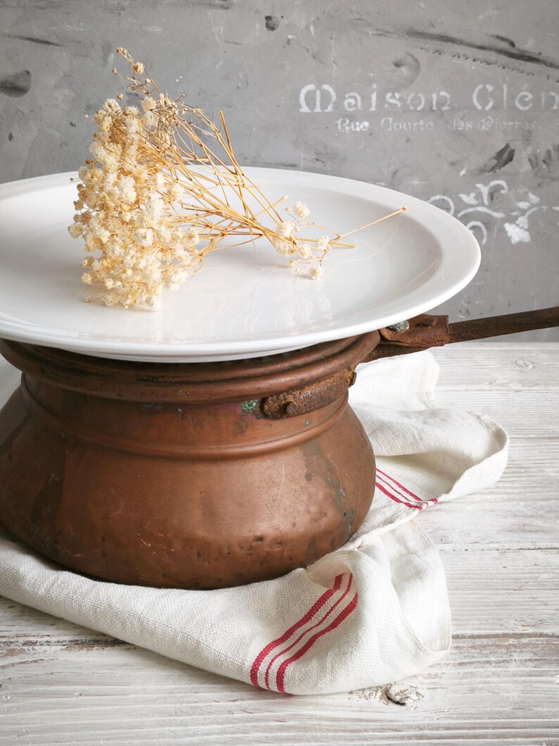Puede incluir: Un plato redondo de cer&aacute;mica blanca descansa sobre una olla de cobre desgastada con un mango de metal. Un peque&ntilde;o racimo de flores secas de paniculata de color crema yace sobre la superficie del plato. La olla de cobre muestra oxidaci&oacute;n.