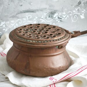 Puede incluir: Una olla de cobre vintage con cuerpo redondeado y tapa plana circular que presenta una rejilla de peque&ntilde;as perforaciones circulares espaciadas uniformemente. La tapa tiene un borde decorativo de cuentas y un mango de metal en el lateral.