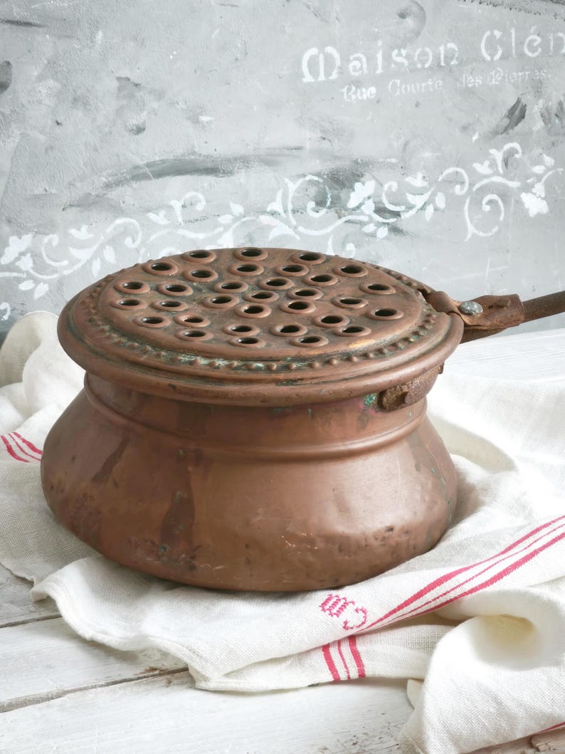 Puede incluir: Una olla de cobre vintage con cuerpo redondeado y tapa plana circular que presenta una rejilla de peque&ntilde;as perforaciones circulares espaciadas uniformemente. La tapa tiene un borde decorativo de cuentas y un mango de metal en el lateral.