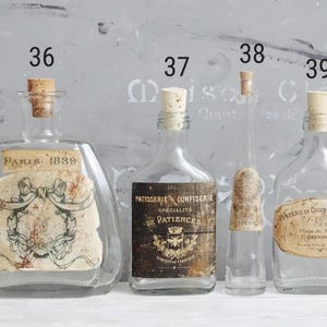 Puede incluir: Cuatro botellas de vidrio transparente con tapones de corcho, numeradas 36, 37, 38 y 39-1. La botella 36 tiene una etiqueta Paris 1889 con un lazo. La 37 tiene una etiqueta Patisserie Confiserie. La 38 es delgada. La 39-1 tiene una etiqueta Fonderie.