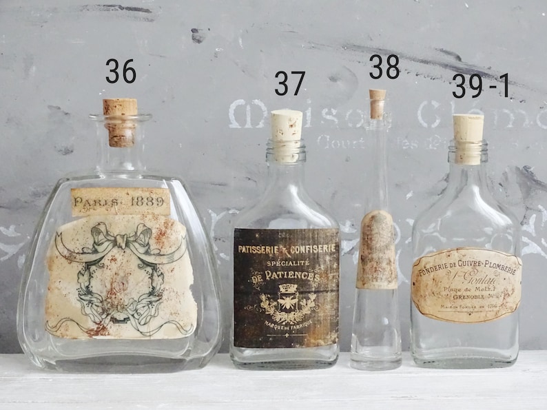 Puede incluir: Cuatro botellas de vidrio transparente con tapones de corcho, numeradas 36, 37, 38 y 39-1. La botella 36 tiene una etiqueta Paris 1889 con un lazo. La 37 tiene una etiqueta Patisserie Confiserie. La 38 es delgada. La 39-1 tiene una etiqueta Fonderie.